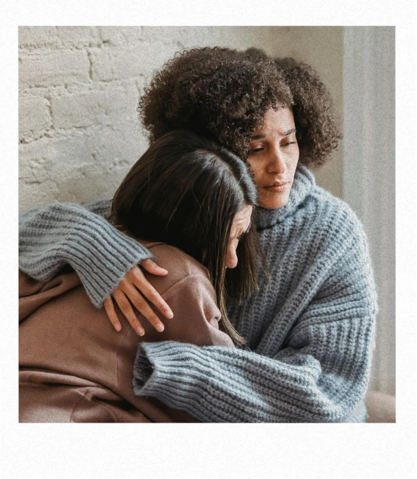 A woman in a chunky light-blue sweater gently hugs and comforts another woman resting her head on her shoulder while they sit against a light brick wall, conveying care and emotional support.