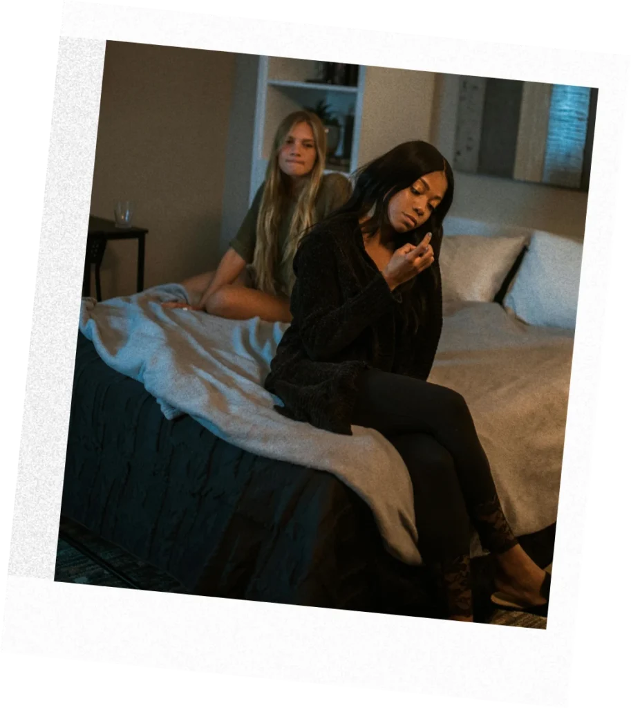 Two women sit on a bed in a dimly lit bedroom, one near the pillows and the other at the edge looking thoughtful
