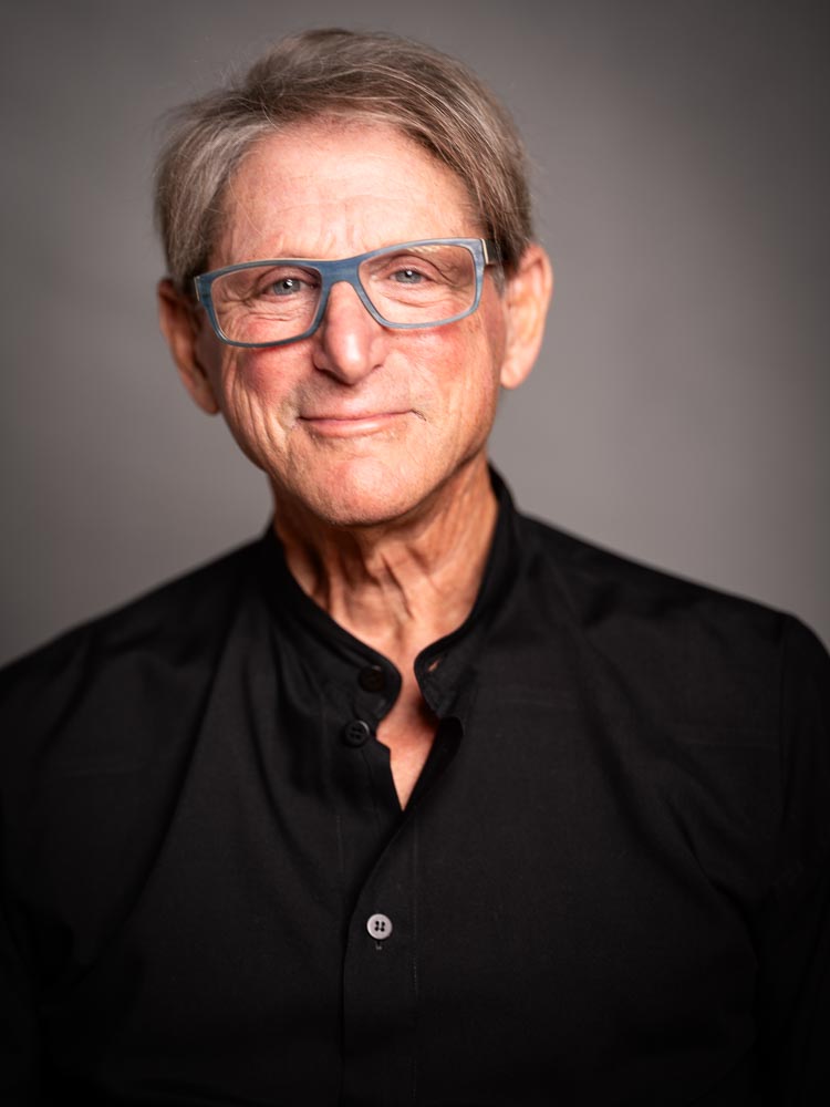 Terry Real looks at the camera with a welcoming and gentle gaze, wearing glasses and a black button-down shirt in a studio photo.