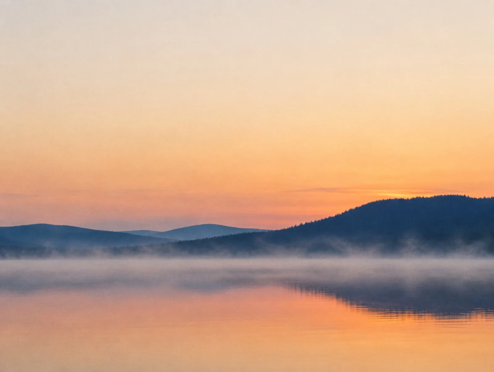 Mist drifting over a still lake at sunrise with wooded hills behind